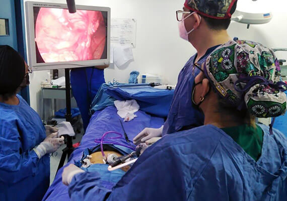 Dra. Irasema Salgado Falcón junto a colegas con bata azul y gorros, viendo laparascopia en pantalla
