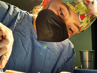 Dra. Irasema Salgado Falcón con bata azul, gorro y cubrebocas negro, haciendo una incisión para operar